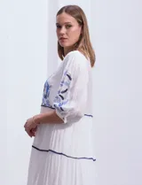 Vestido midi blanco con bordados florales azules en el escote, mangas y cintura. Tiene cuello en V, mangas 3/4 con botón y falda escalonada con detalles de ribetes azules.