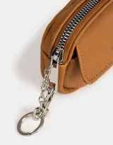 Monedero color marrón camel de suede con bolsillo frontal con solapa y broche con botón para cerrado. Tiene cierre superior con mosquetón y tag en relieve con la inscripción "Girls Club".