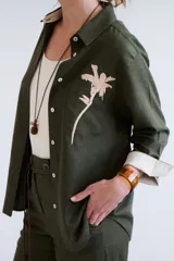 Camisa verde militar de manga larga con puños color crema, cuello clásico y cierre frontal con botones. Tiene un bolsillo en el pecho con bordado de una palmera color crema.
