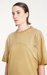 Remera oversize de algodón, color mostaza con efecto batik artesanal. Presenta costuras decorativas arqueadas que unen el cuello con los hombros y el torso.