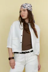 Chaqueta color crudo de lino con cuello redondo, manga tres cuartos, cierre frontal con botones y bolsillos de parche. Detalle de costuras en contraste color marrón.