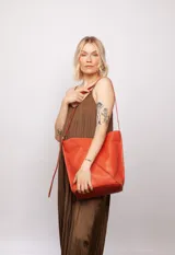 Bolso tipo bucket de cuero color naranja, con costuras externas, cierre con broche imán, correa regulable, dos bolsillos internos (uno con cierre) e interior de algodón.