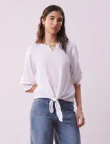 Blusa blanca de tejido texturado con cuello redondo, escote en V con aplique metálico y mangas 3/4 con puños elásticos. El ruedo se ata con un nudo.