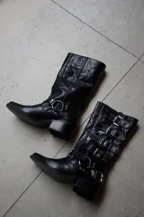 Bota de cuero vacuno color rojo, de caña alta y punta fina, con detalles de tiras con hebillas plateadas. El interior está forrado en cuero vacuno. Tiene un fondo de doble vira de madera.