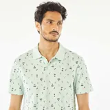 Remera polo verde claro con estampado de palmeras y tablas de surf en negro.