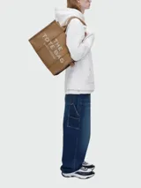 Bolso tote grande de tela negra con asas de mano y hombro. Presenta el texto "THE TOTE BAG MARC JACOBS" en color blanco en el frente. Los bordes laterales tienen una cinta blanca con texto repetido.