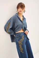 Camisa de denim azul de corte amplio, manga larga, cuello camisero y bolsillos en el delantero. Presenta bordados dorados de mostacillas, lentejuelas y canutillos en mangas y parte inferior de un lateral del pantalón.