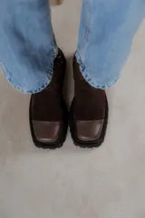 Bota corta de cuero gamuzado color marrón chocolate, con punta cuadrada y suela track. Presenta una correa con hebilla desmontable y cierre lateral con cremallera.