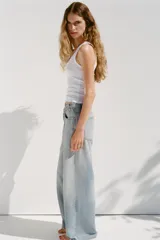 Pantalón de jean celeste de tiro alto, corte wide leg y largo hasta el tobillo. Presenta un diseño asimétrico en la cintura con cierre frontal cruzado con cremallera y botones metálicos.