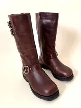 Bota de caña media de cuero vacuno color marrón oscuro, con punta cuadrada, cierre lateral y suela track negra. Presenta detalles de hebillas metálicas plateadas en la caña y en la parte inferior del empeine.