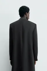 Levita negra de corte relajado confeccionada en tejido con lana. Presenta cuello sin solapa, mangas largas con puños abotonados, bolsillo de vivo en el pecho y bolsillos con solapa en la cadera. Incluye bolsillos interiores, abertura en el centro de la espalda y cierre frontal con botonadura.