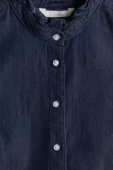 Camisa de denim azul oscuro con cuello mao y volados en cuello y puños. Tiene mangas largas abullonadas con botón en los puños, botonadura frontal y bajo redondeado.