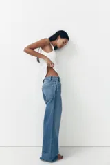 Pantalón de jean celeste de tiro alto, corte wide leg y largo hasta el tobillo. Presenta un diseño asimétrico en la cintura con cierre frontal cruzado con cremallera y botones metálicos.