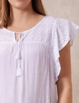 Blusa blanca de gasa con broderie, cuello a la base, escote en V y lazo de ajuste. Delantera broderie con corte y frunces. Manga corta volado de broderie. Espalda con corte y frunces.