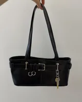 Cartera de hombro negra, de forma rectangular y acabado liso. Presenta detalles de hebillas plateadas y tiras decorativas en el frente, junto con un dije colgante en forma de llave dorada.