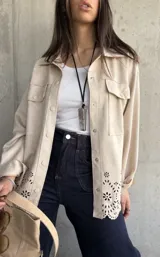 Sobrecamisa color beige de gamuza sintética, corte oversize, con cuello camisero, cierre frontal con botones a presión, bolsillos de parche en el pecho y detalle calado con motivos florales en el ruedo.