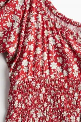 Blusa roja con estampado de flores blancas, escote cuadrado con cordón ajustable, mangas cortas abullonadas con elástico en hombros y puños, cintura fruncida y volados en mangas y bajo.