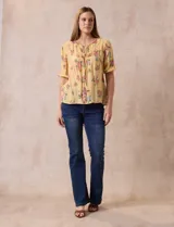 Blusa amarilla estampada con flores rosadas, de gasa, con cuello a la base, escote en V plisado y lazo de ajuste. Delantera con detalles en puntilla y mangas 3/4 con puño fruncido.