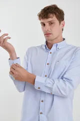 Camisa celeste de tejido texturado oxford, corte clásico, con cuello abotonado, mangas largas con puños ajustables, bolsillo en el pecho y cierre frontal con botones de madera.