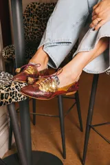 Zapato blucher de cuero con estampado animal print en tonos marrones y dorados, con cordones finos color borgoña y detalle de flor en el extremo del cordón.