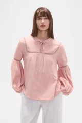 Blusa holgada de algodón color rosa pálido, con escote redondo y lazo ajustable en el centro. Presenta detalles de encaje calado alrededor del escote y en los puños de las mangas largas abullonadas.