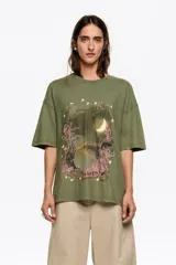 Camiseta de manga corta, corte oversize y escote caja, color verde militar. Presenta un estampado frontal con temática nocturna, incluyendo una luna creciente, árboles y texto en tonos rosados.