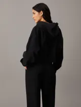Buzo negro cropped con capucha y cierre frontal de cremallera metálica. Tiene bolsillos de parche en la cintura y logo de Calvin Klein bordado en el pecho.