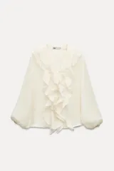Camisa color crudo confeccionada en tejido de lyocell, con escote en pico, mangas largas abullonadas con elástico en los puños y volantes frontales.