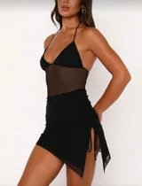 Vestido negro de corte ajustado, con escote halter y sección central de malla transparente. Incluye una minifalda negra superpuesta con frunces laterales ajustables mediante lazos.
