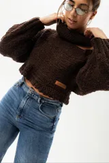 Sweater tejido en lana merino color marrón oscuro, con cuello alto y un hombro descubierto con tira ajustable con hebilla metálica.