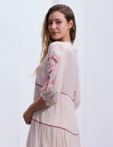 Vestido midi blanco con bordados florales azules en el escote, mangas y cintura. Tiene cuello en V, mangas 3/4 con botón y falda escalonada con detalles de ribetes azules.