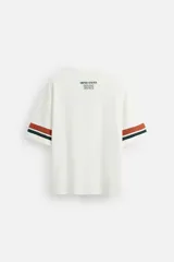 Camiseta blanca de corte holgado con cuello redondo y cierre frontal de botonadura oculta. Tiene mangas cortas con rayas horizontales en marrón, verde y azul. Presenta un parche bordado en el pecho y texto bordado en el frente.