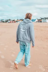 Hoodie gris con capucha, cordón ajustable y bolsillo tipo canguro. Estampado en la espalda con el logo de la marca: un círculo con el texto "The Glassy Club Since 11", una flor, una tabla de surf y una bandera a cuadros.