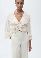 Blusa color crema con cuello en V, volados y detalles de encaje. Tiene mangas largas con puños elásticos y lazos.