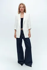Blazer blanco con cuello solapa y hombreras. Manga por debajo del codo acabada en vuelta con trabilla y botón dorado. Bolsillos delanteros con solapa.