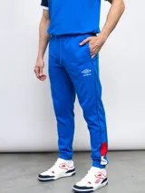 Pantalón de jogging azul marino con puños ajustados, cintura elástica con cordón y bolsillos laterales. Presenta una franja blanca con detalles en rojo a lo largo de la pierna, el escudo del Club Nacional de Football y el logo de Umbro.