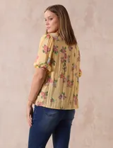 Blusa amarilla estampada con flores rosadas, de gasa, con cuello a la base, escote en V plisado y lazo de ajuste. Delantera con detalles en puntilla y mangas 3/4 con puño fruncido.