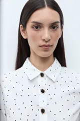 Blusa blanca con estampado de pequeños lunares negros, cuello camisero y botones frontales oscuros. Mangas tres cuartos con ligero vuelo en el puño.