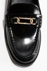 Mocasines negros de charol con suela gruesa y costura tradicional en la puntera. Presentan una tira decorativa con una hebilla metálica dorada con diseño de eslabones entrelazados.