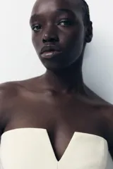 Top blanco de escote recto con detalle de pieza metálica en forma de V forrada y hombros descubiertos. Tiene falsos bolsillos con solapa en el delantero, forro interior combinado y cierre lateral con cremallera oculta en la costura.