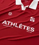 Remera de manga corta estilo polo, color rojo, con cuello y puños en contraste blanco. Presenta un corte oversized con hombros caídos y la palabra "ATHLÈTES" estampada en blanco en el frente. Incluye pequeños gráficos blancos a cada lado del pecho.