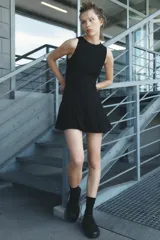 Vestido corto negro sin mangas, con cuello redondo y falda con tablas. Cierre trasero con cremallera oculta.