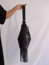 Bolso tipo bandolera de cuero negro con flecos y tachas plateadas. Tiene correa regulable, forro interior y bolsillo interno.