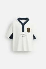 Camiseta blanca de corte relaxed fit, con cuello pico y cierre frontal de botonadura oculta. Presenta mangas cortas y detalles color block en azul marino en cuello y mangas. Incluye bordados y parche con motivos deportivos en el frente.