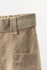 Bermuda color beige de lino, con cintura elástica, trabillas y cinturón con hebilla metálica. Presenta bolsillos de parche laterales y traseros.