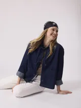 Chaqueta de jean azul oscuro con cierre frontal con lazos, mangas 3/4 con puños doblados, bolsillos de parche en el frente y corte holgado.