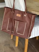 Bolso tipo tote de cuero color marrón con costuras blancas en contraste, bolsillo externo, doble asa de hombro y cierre superior con cremallera.