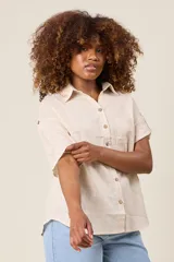 Camisa color beige claro, de corte clásico, con cuello, botones frontales, bolsillo en el pecho y mangas tres cuartos con presilla para sujetar.