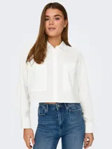 Camisa blanca de corte cropped, con cuello clásico, mangas largas con puños abotonados y dos bolsillos de parche en el pecho.