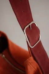 Bolso de hombro tipo hobo de cuero gamuzado color camel, con correa ancha ajustable con hebilla metálica plateada. La correa presenta un estampado de vaca (cow print) en tonos blanco y marrón.